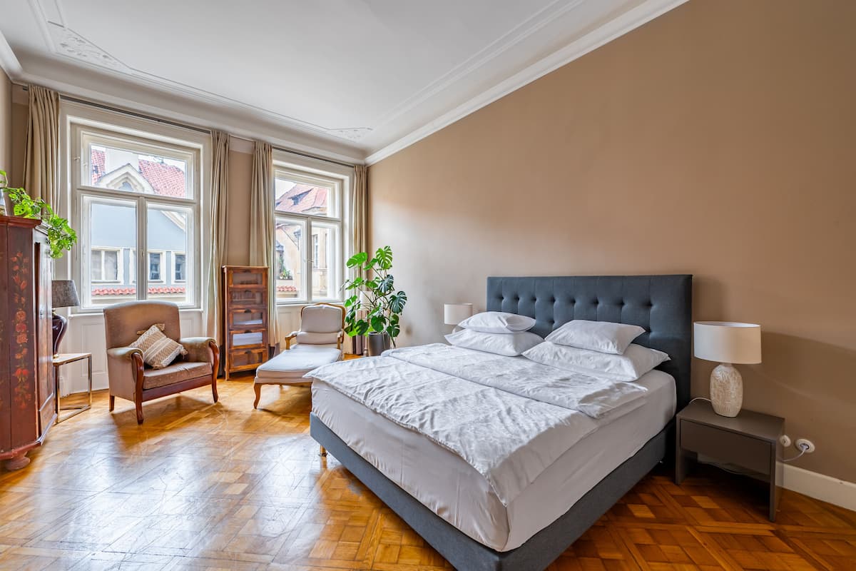 A bedroom with a large bed, a nightstand, a chair, a desk, and two windows with curtains.