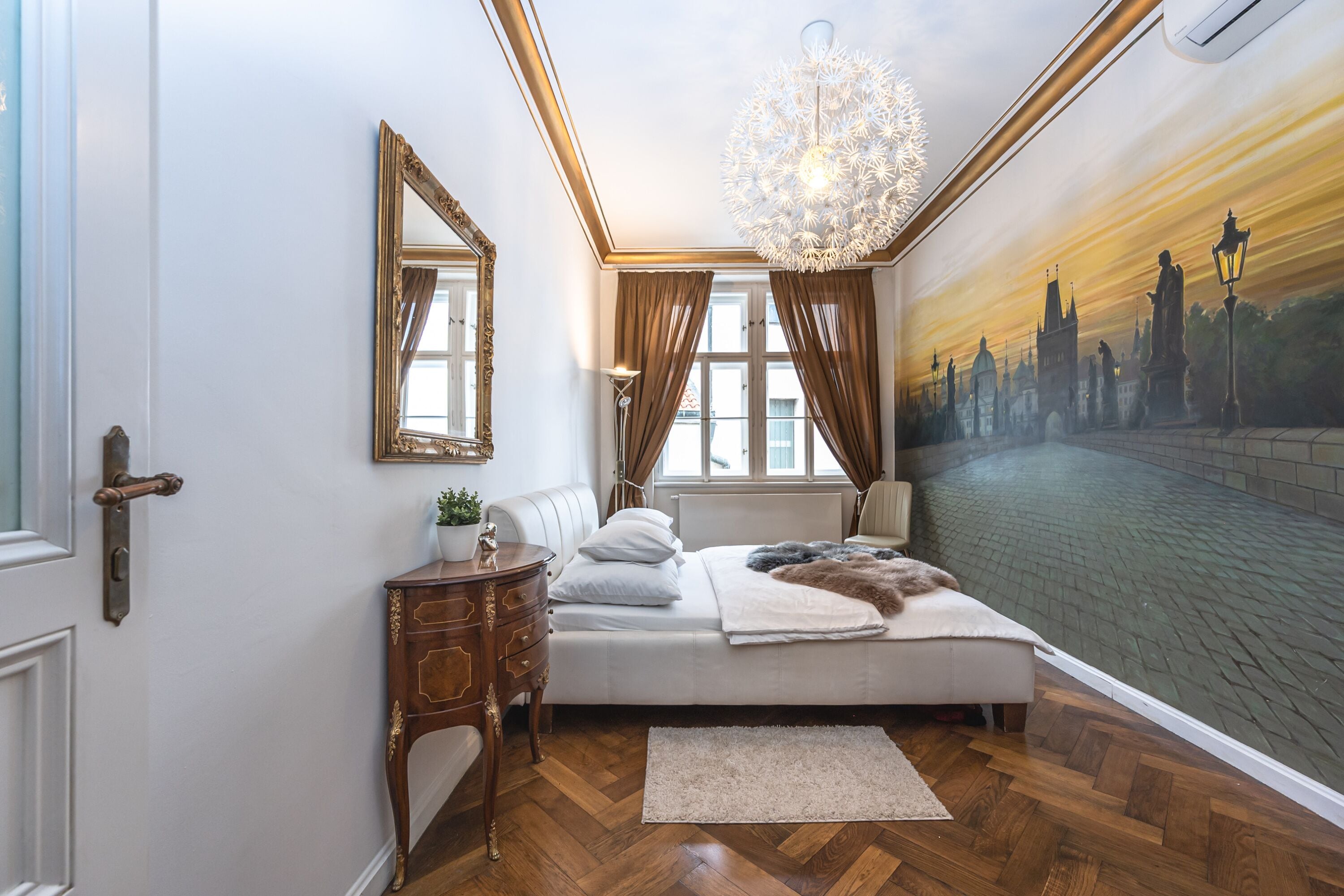 Une chambre à coucher avec un grand lit, une table de chevet en bois, un miroir et une fresque murale représentant un paysage urbain.