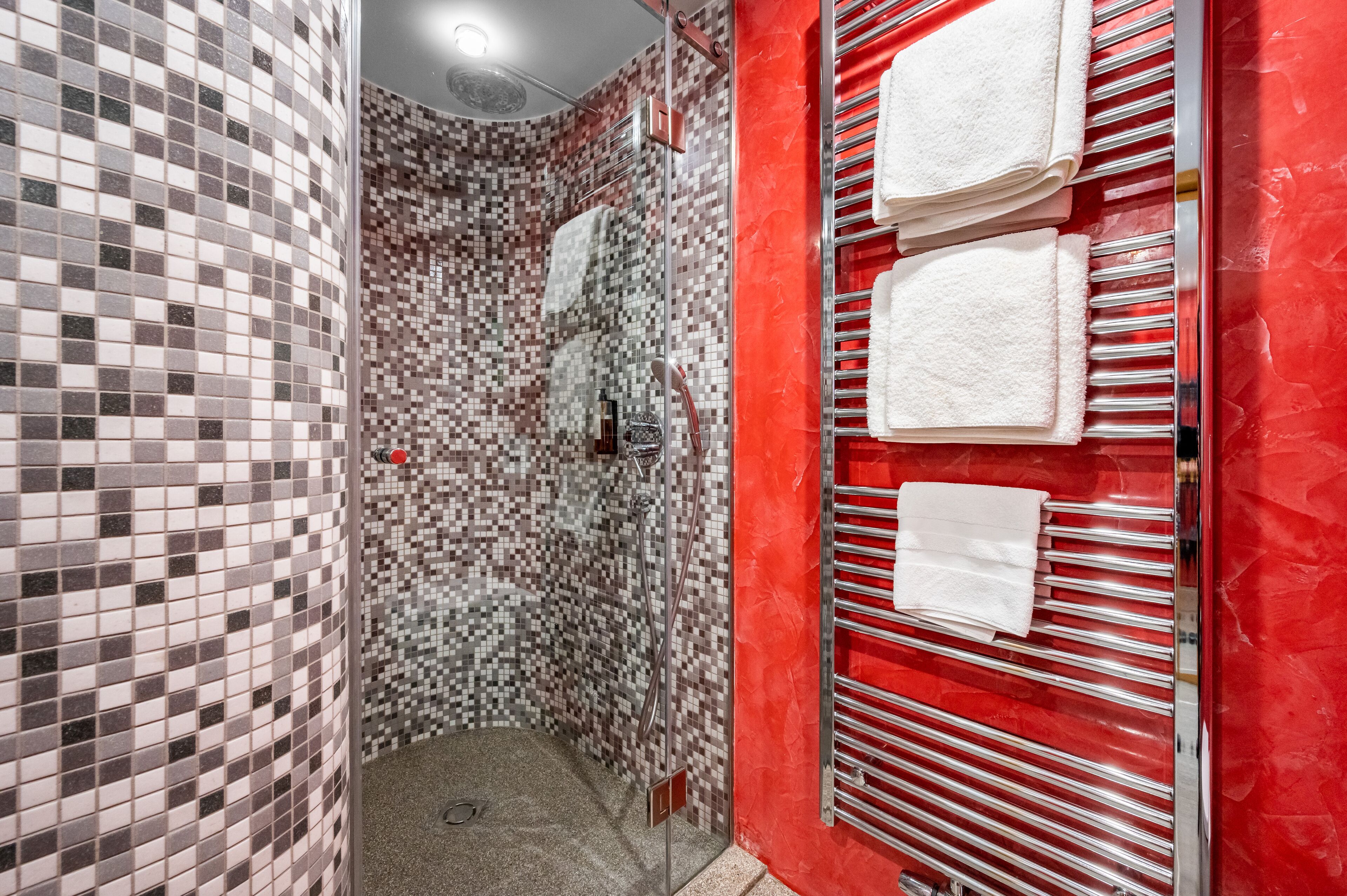 Un baño con zona de ducha azulejada, una pared roja y un perchero con varias toallas.