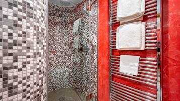 Un baño con zona de ducha azulejada, una pared roja y un perchero con varias toallas.