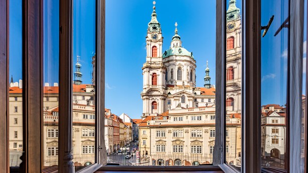 Vista a través de una ventana que muestra un edificio con una iglesia grande y ornamentada, con dos torres y una cúpula, rodeada de otros edificios históricos.