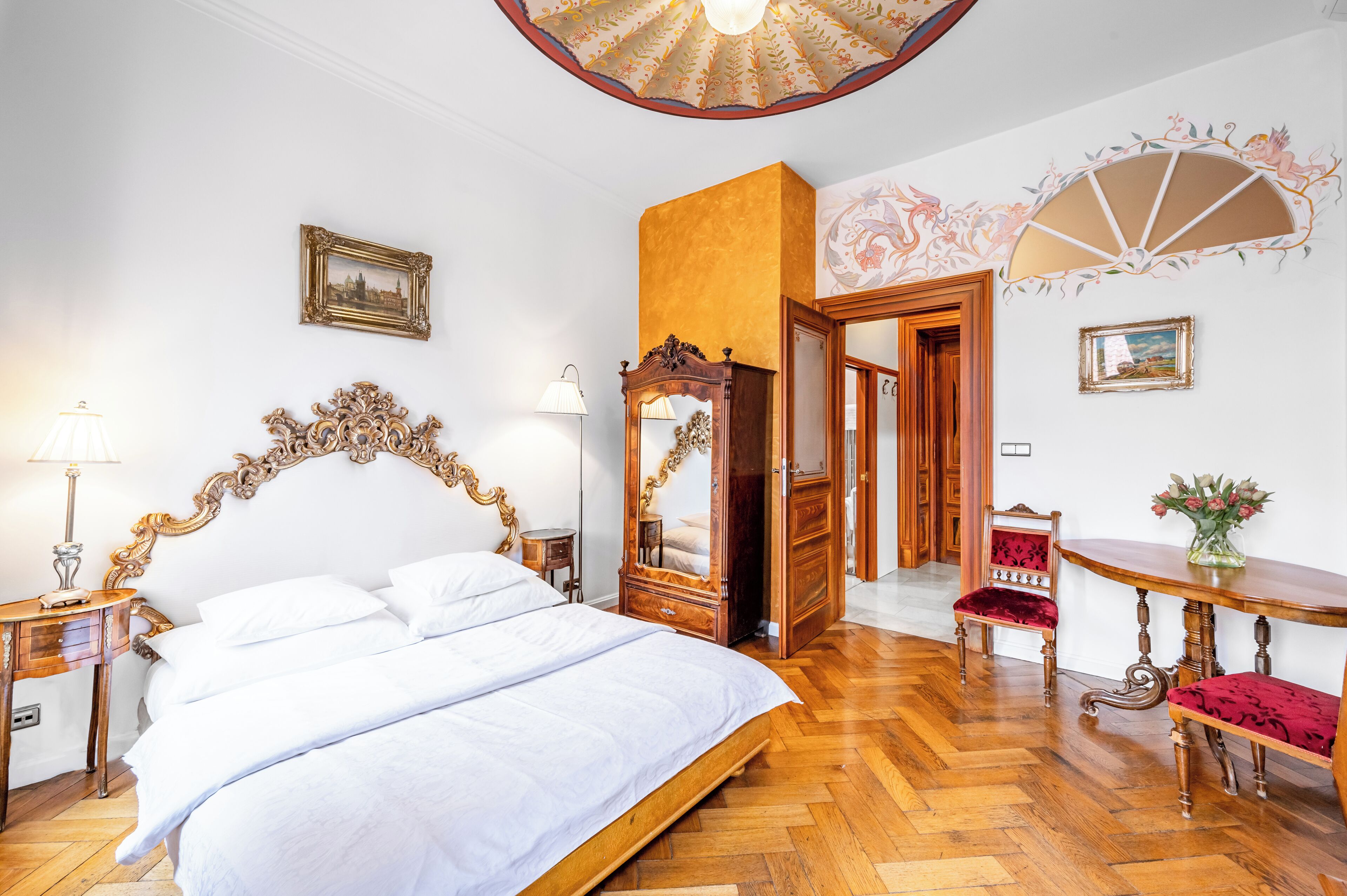 A bedroom with a large bed, a wooden headboard, a mirror, a red chair, a wooden table with a vase, and a door leading to another room.