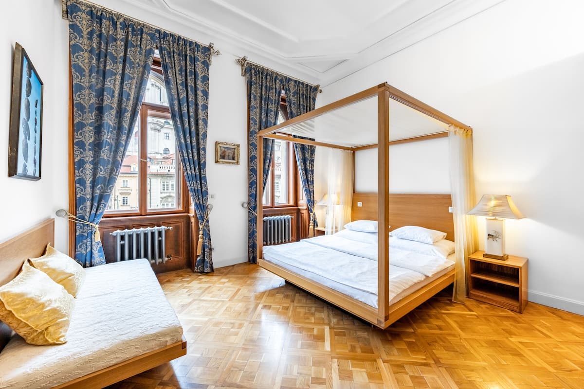 A room with a four-poster bed, a smaller bed, wooden flooring, and a view of buildings through large windows with patterned curtains.