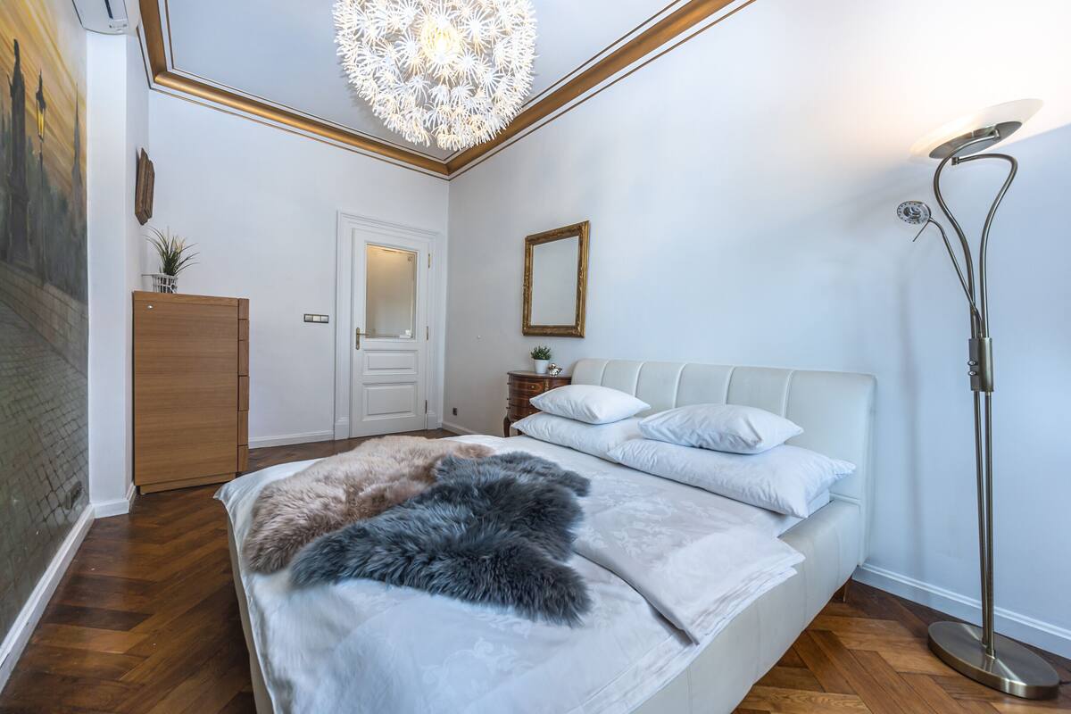 A bedroom with a large bed, a wooden wardrobe, a nightstand, a mirror, and a floor lamp.