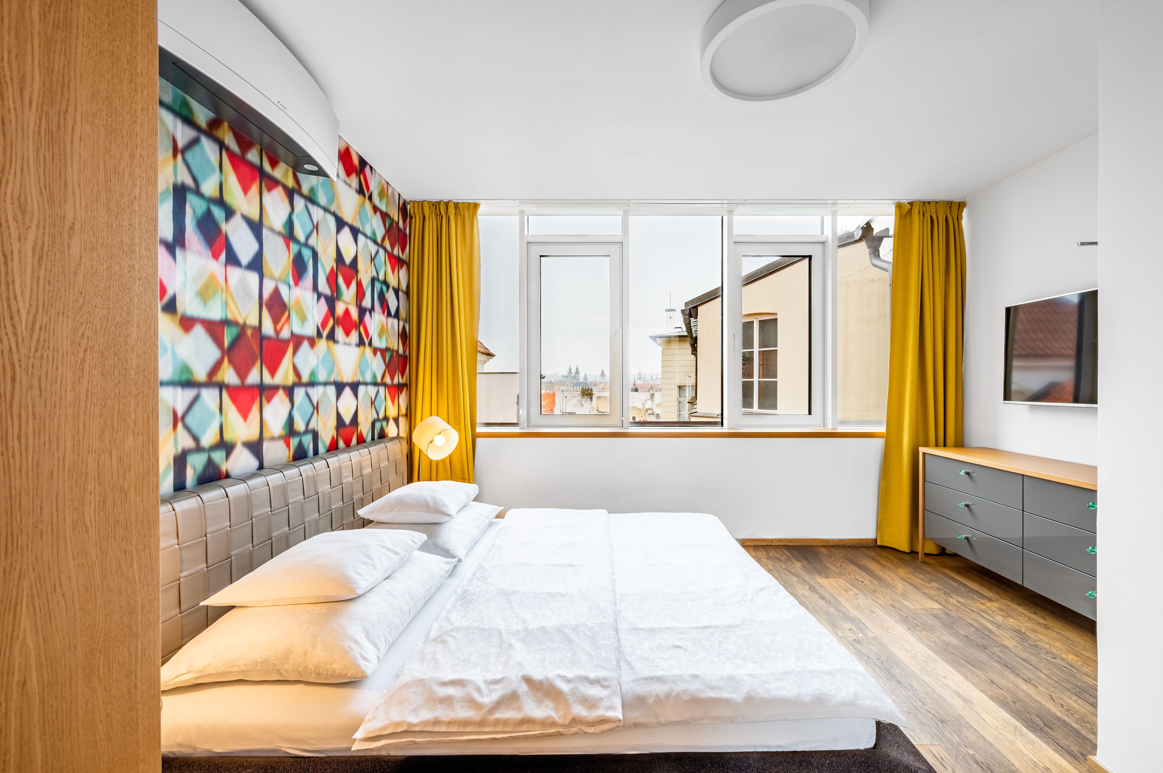 A modern hotel room with a large bed, a colorful geometric wall art, a large window with a view of the city, and a wooden headboard.