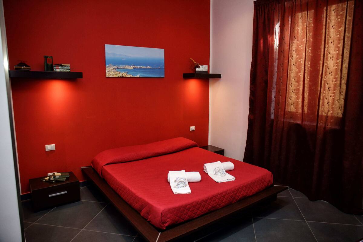 Habitación de hotel con pared roja, cama con colcha roja, dos toallas sobre la cama, mesita de noche con lámpara, estante con libros y una foto, y ventana con cortinas.