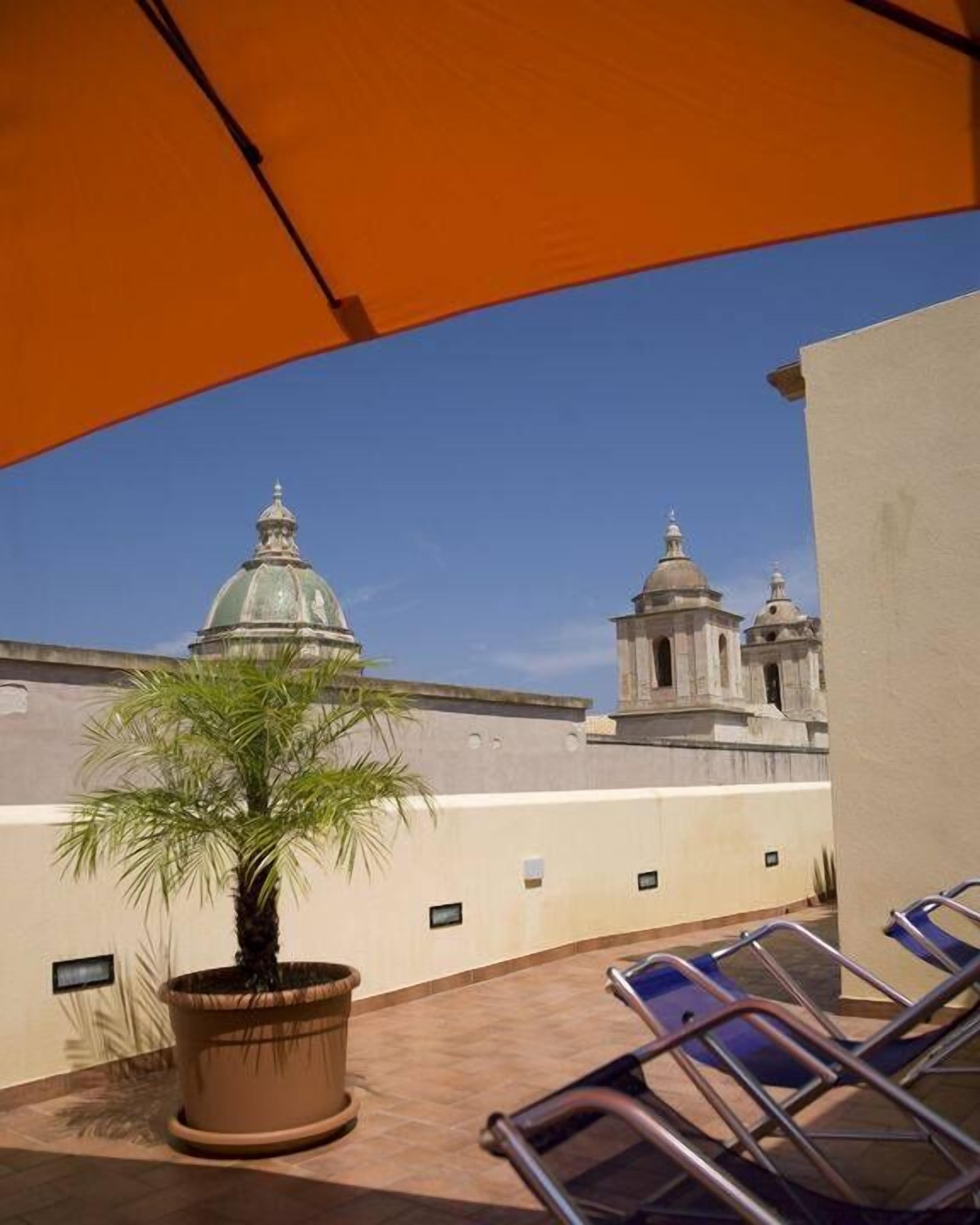 Eine Dachterrasse mit einer Palme, Liegestühlen und Blick auf ein historisches Gebäude mit Kuppeln.