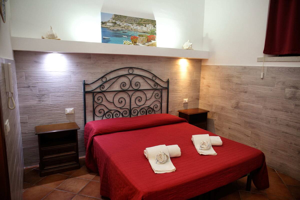 Una habitación de hotel con una cama, dos mesitas de noche, un cuadro en la pared y una cortina roja.