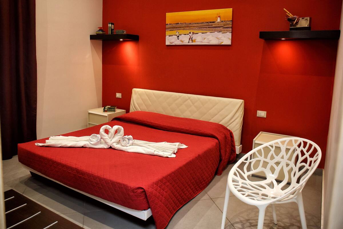 Una habitación de hotel con una colcha roja, una silla blanca y un cuadro en la pared.