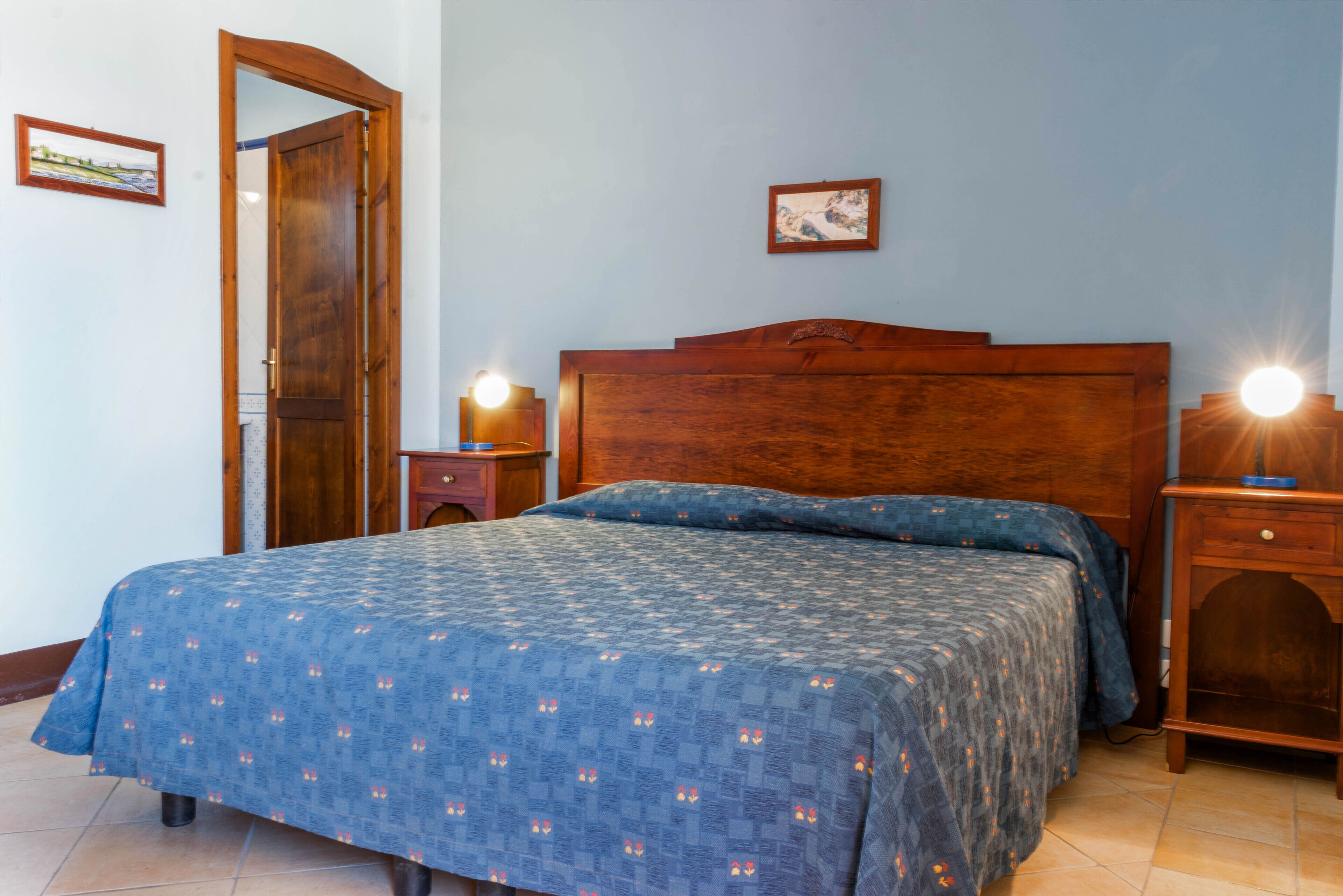Een slaapkamer met een houten hoofdbord, een bed met een blauw beddegoed met patroon, twee nachtkastjes met lampen, en een deur.