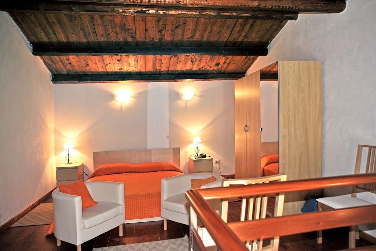 A bedroom with a wooden ceiling, a bed with orange bedding, two bedside tables with lamps, and a wooden chair.