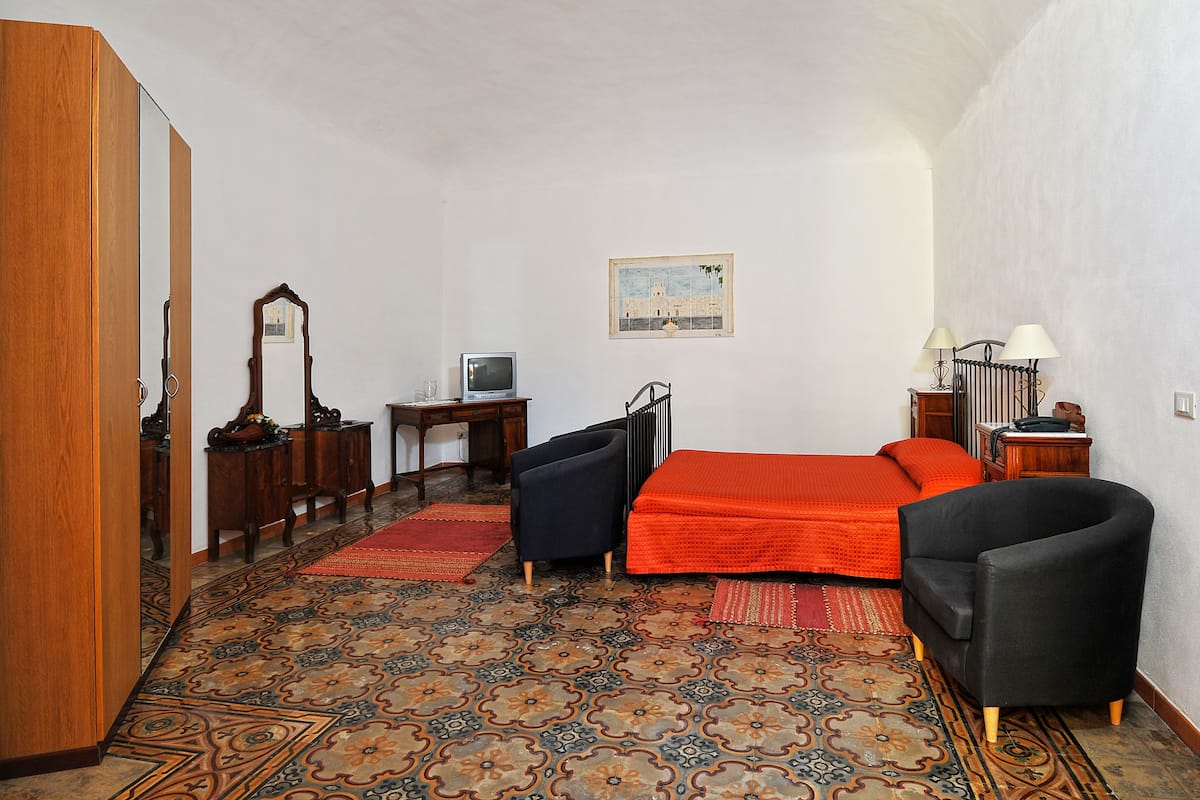 A bedroom with a bed, two chairs, a desk with a computer, a mirror, and a radiator.