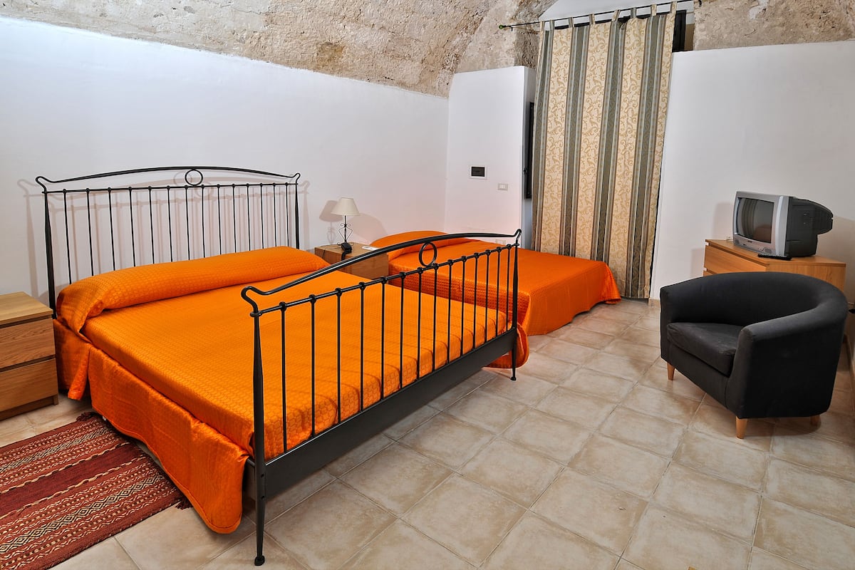 A bedroom with a metal bed frame, orange bedding, a wooden nightstand, a television on a wooden cabinet, and a patterned rug.
