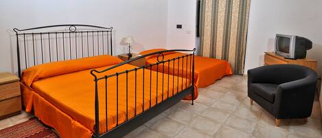 A bedroom with a metal bed frame, orange bedding, a wooden nightstand, a television on a wooden cabinet, and a patterned rug.