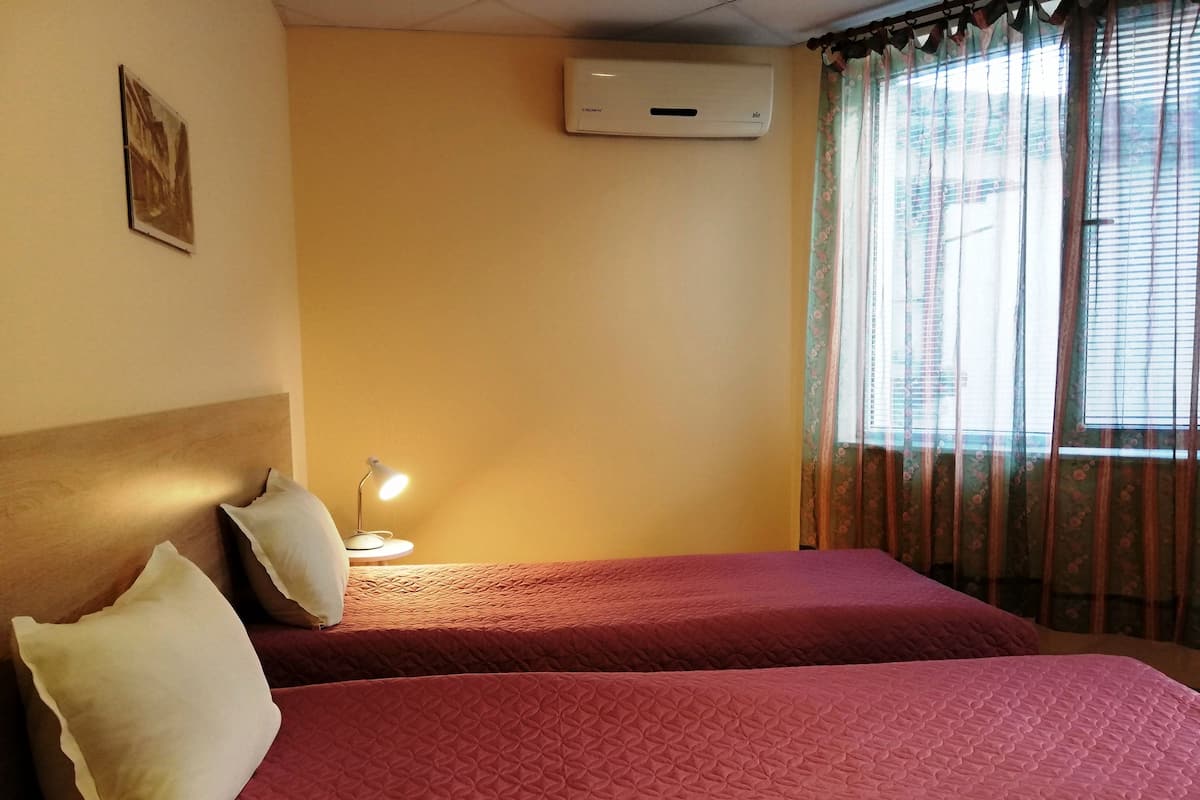 Une chambre d’hôtel avec deux lits, une lampe de chevet, un appareil de climatisation et une fenêtre agrémentée de rideaux à motifs floraux.