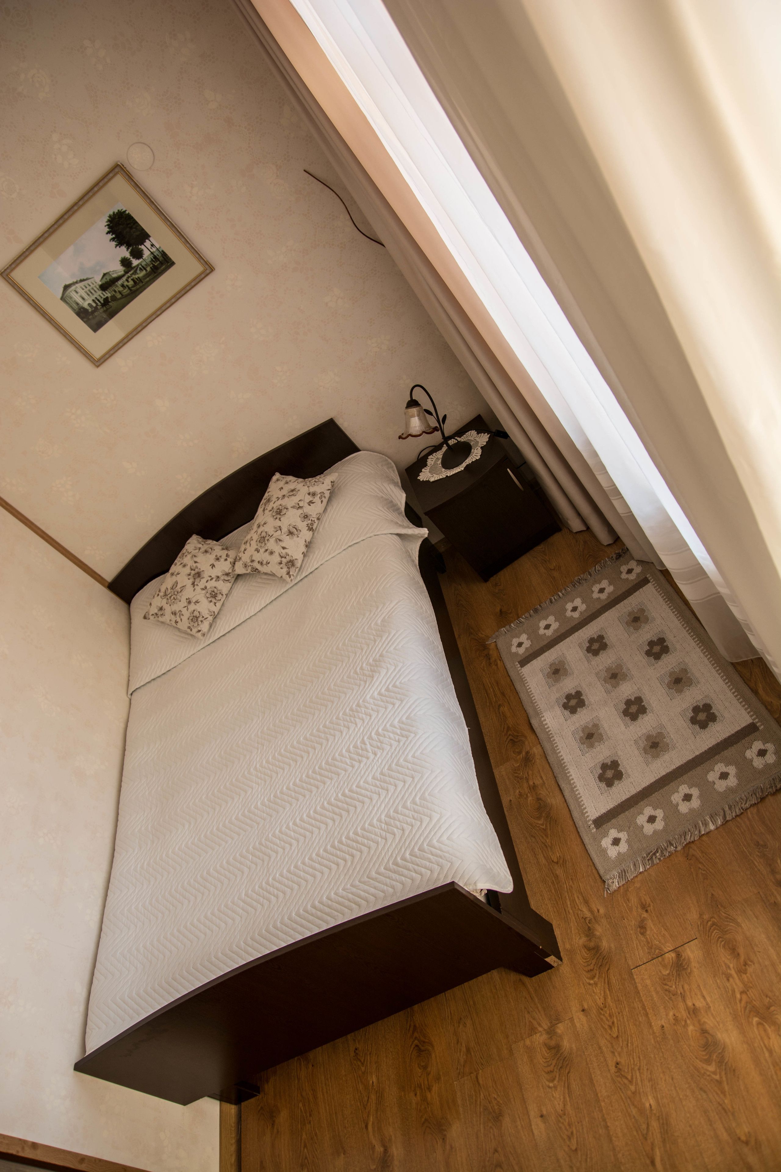 A neatly made bed with a patterned bedspread and pillows, a small bedside table with a lamp, and a rug on a wooden floor.