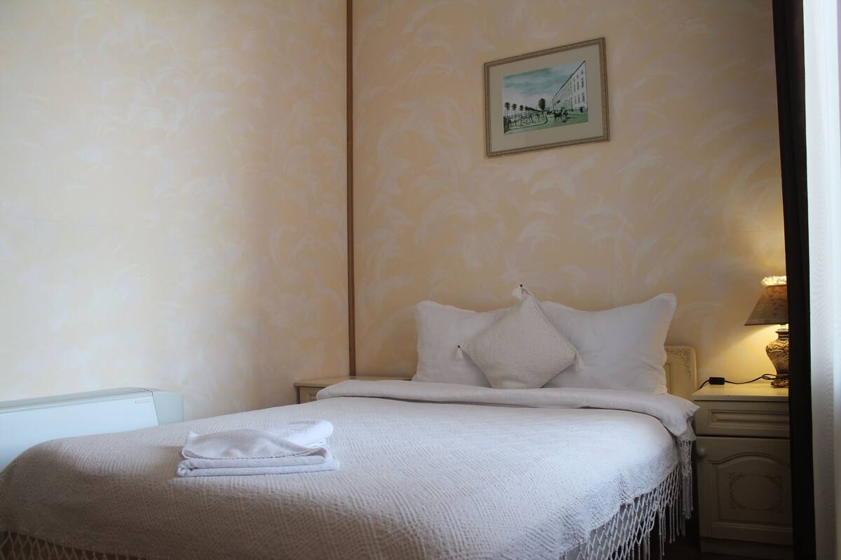 Un lit bien fait, avec un drap blanc et des oreillers, une table de chevet avec une lampe et un tableau encadré au mur.