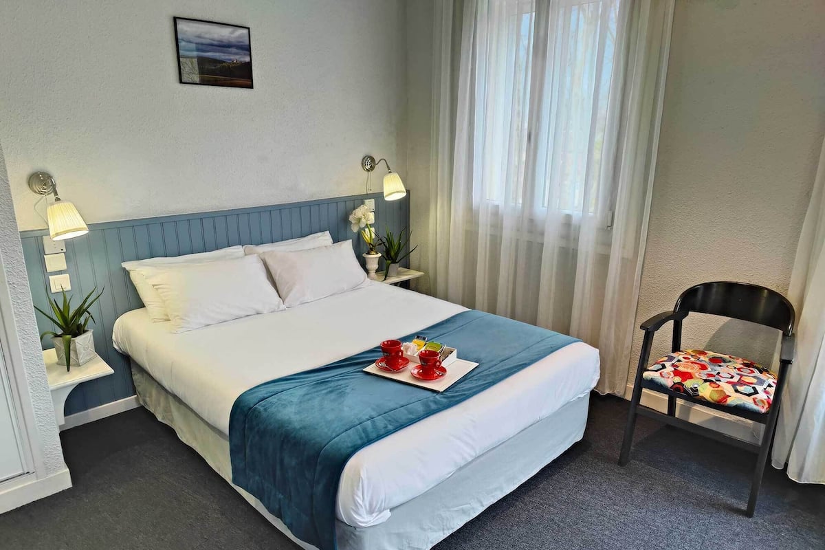 Un lit bien fait, recouvert d’une couvre-lit bleue, avec des oreillers blancs et un plateau sur lequel se trouvent une tasse et une soucoupe rouges. On y voit également une chaise avec un coussin coloré, une plante en pot et une lampe fixée au mur.