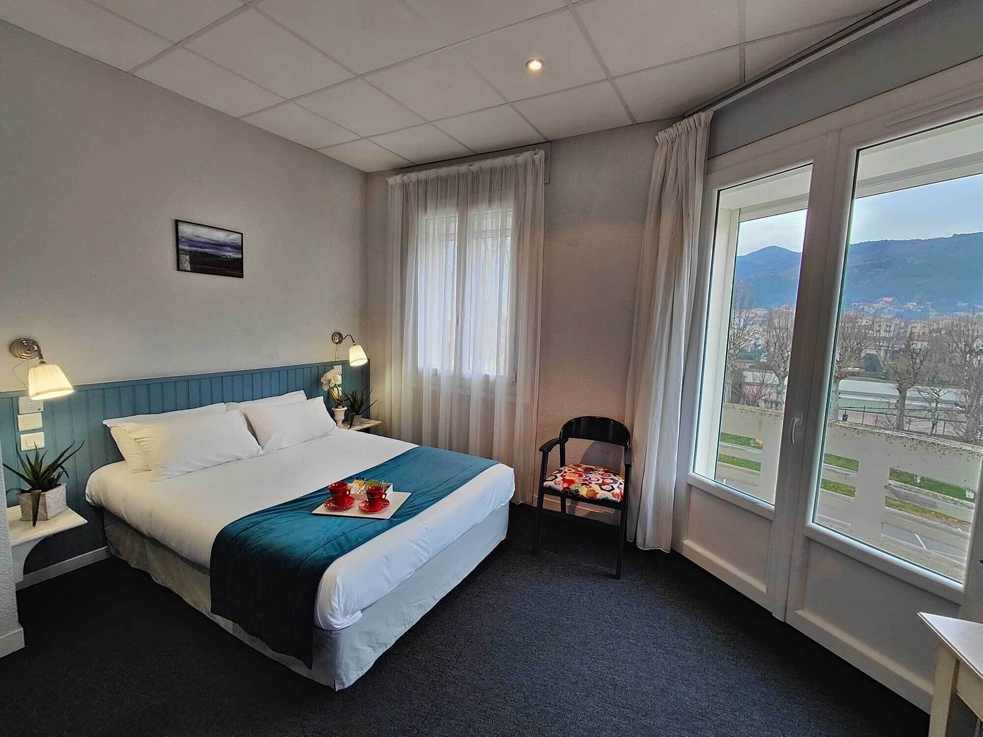 A hotel room with a bed, a chair, a window with a city view, and a nightstand with a lamp.