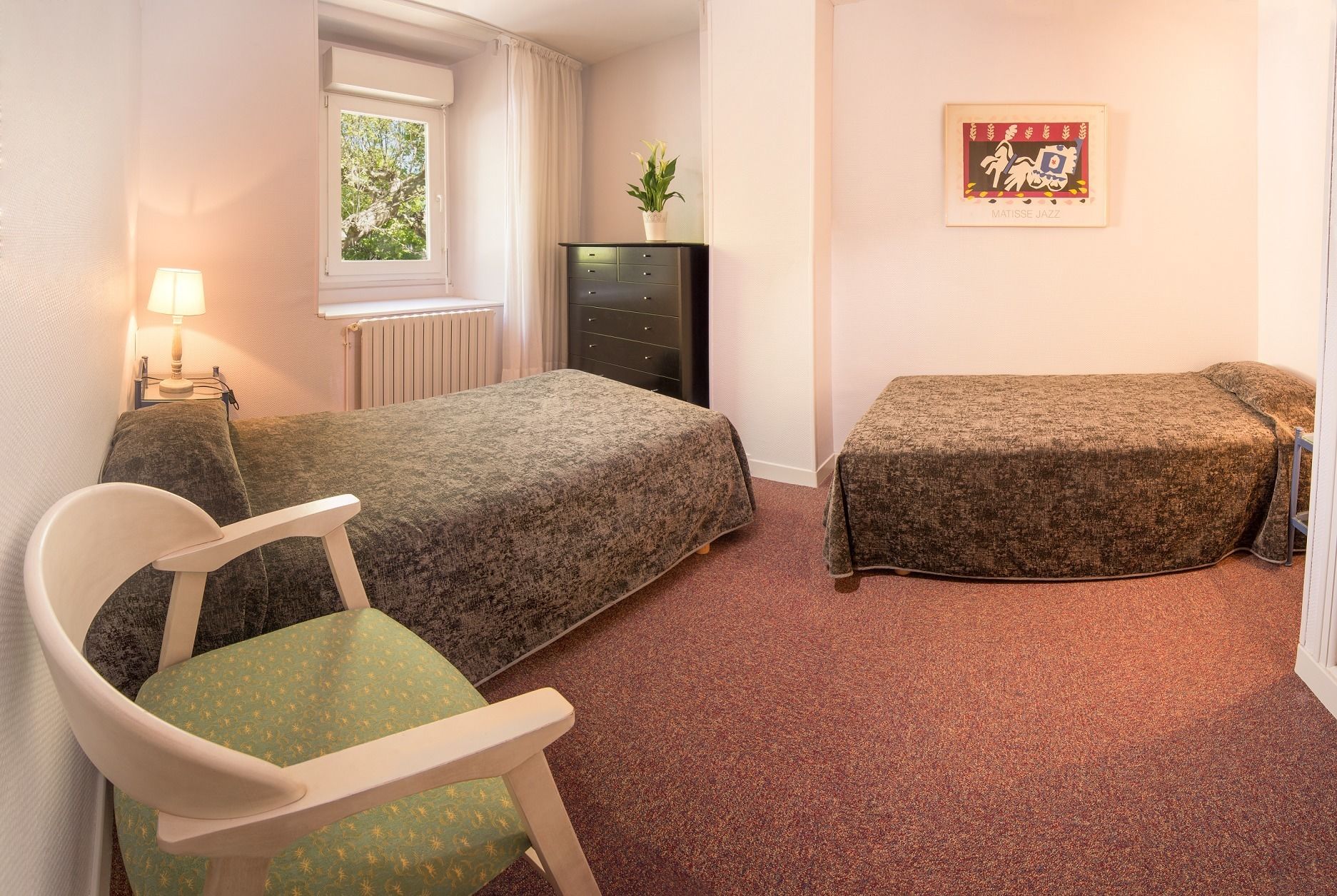 A room with two single beds, a chair, a desk, a radiator, a window, and a framed artwork on the wall.