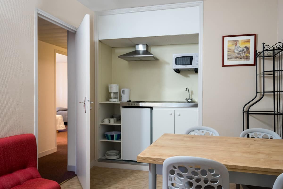 Une cuisine compacte avec des meubles blancs, un four à micro-ondes et une machine à café. On y trouve une table à manger avec des chaises, et une chaise rouge est placée au premier plan.