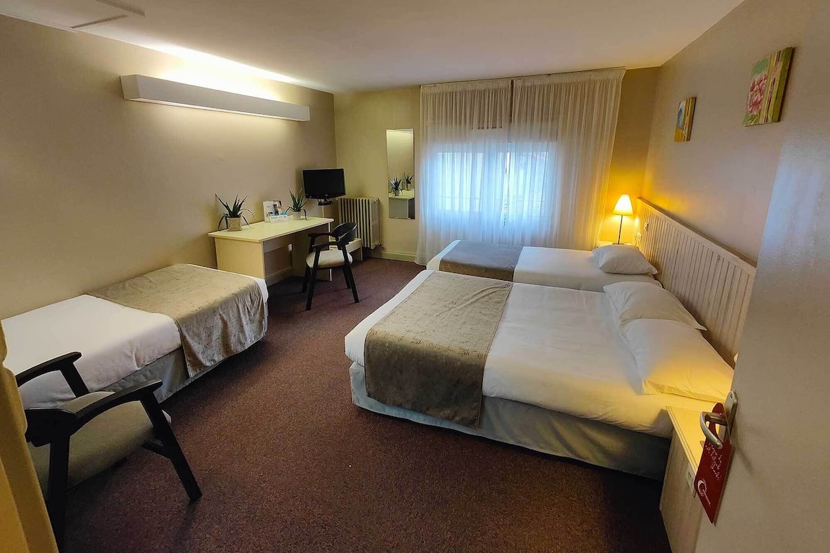 Une chambre d’hôtel avec deux lits simples, un lit double, un bureau avec une chaise, une télévision et une fenêtre avec des rideaux.