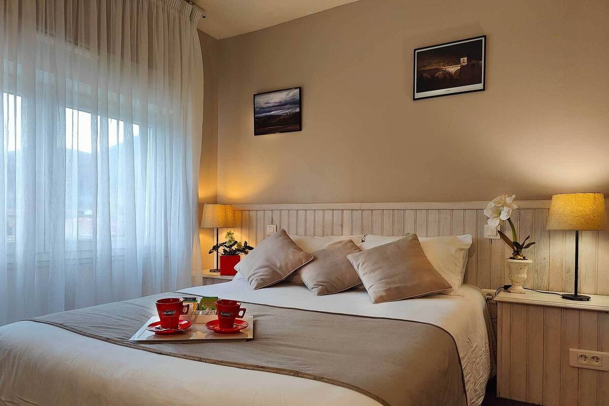 Une chambre d’hôtel avec un lit, deux lampes de chevet, une table de chevet avec une lampe, une fenêtre avec des rideaux légers et un tableau encadré au mur.