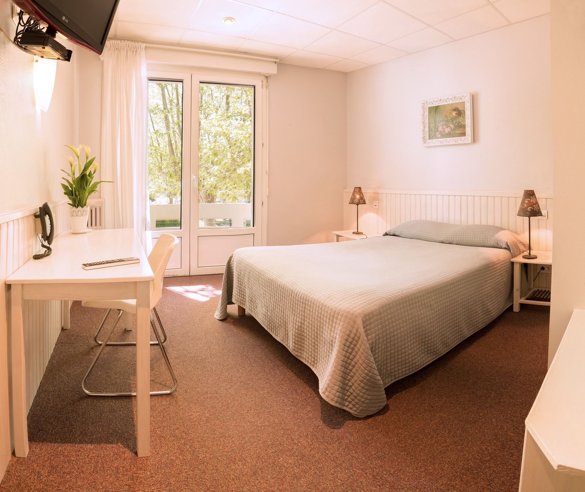 A hotel room with a bed, a desk with a telephone and a plant, a chair, a nightstand with a lamp, and a window with curtains.