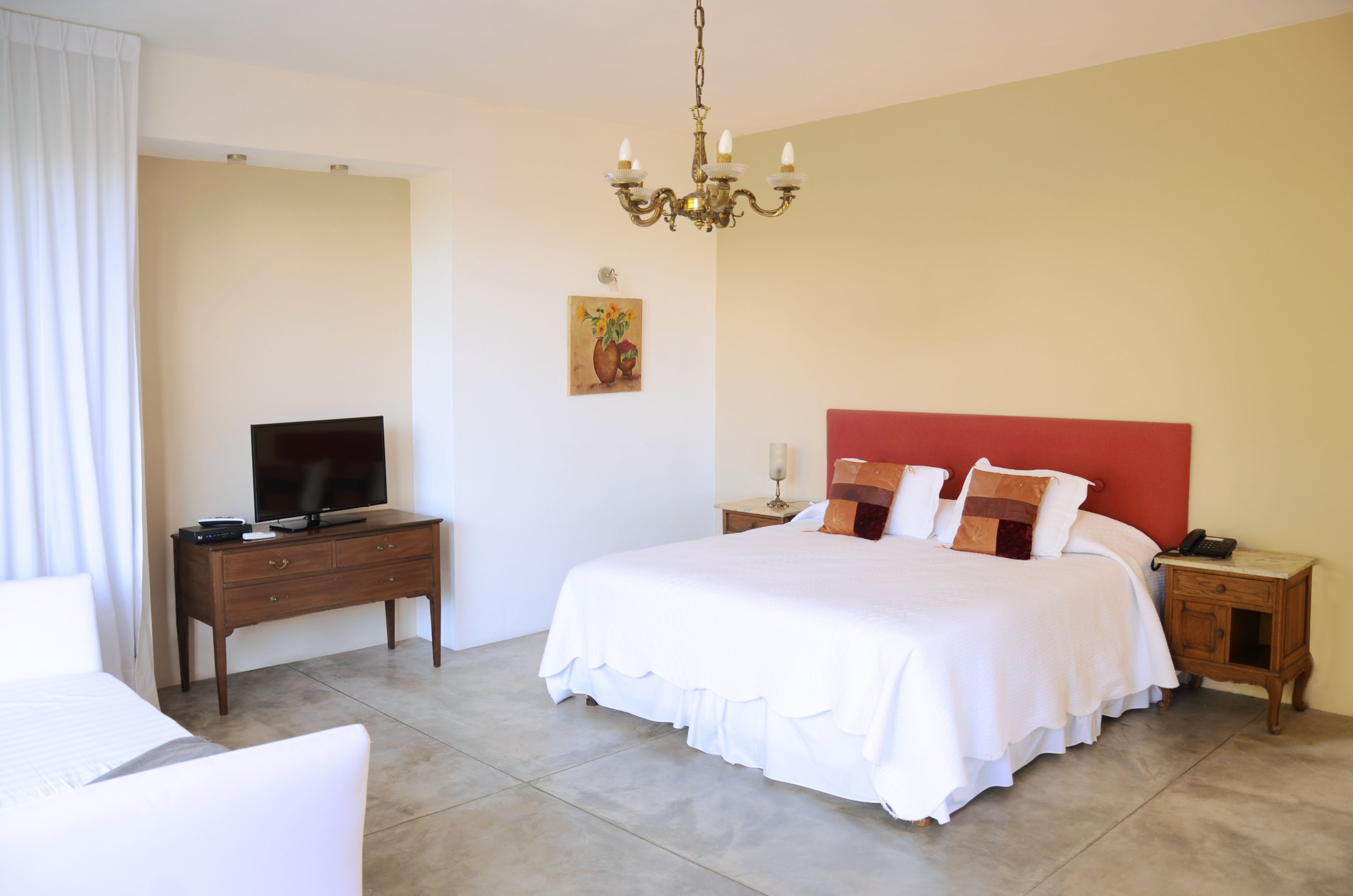 Una habitación de hotel con una cama grande, un televisor sobre un aparador de madera, una mesita de noche con una lámpara y un cuadro en la pared.