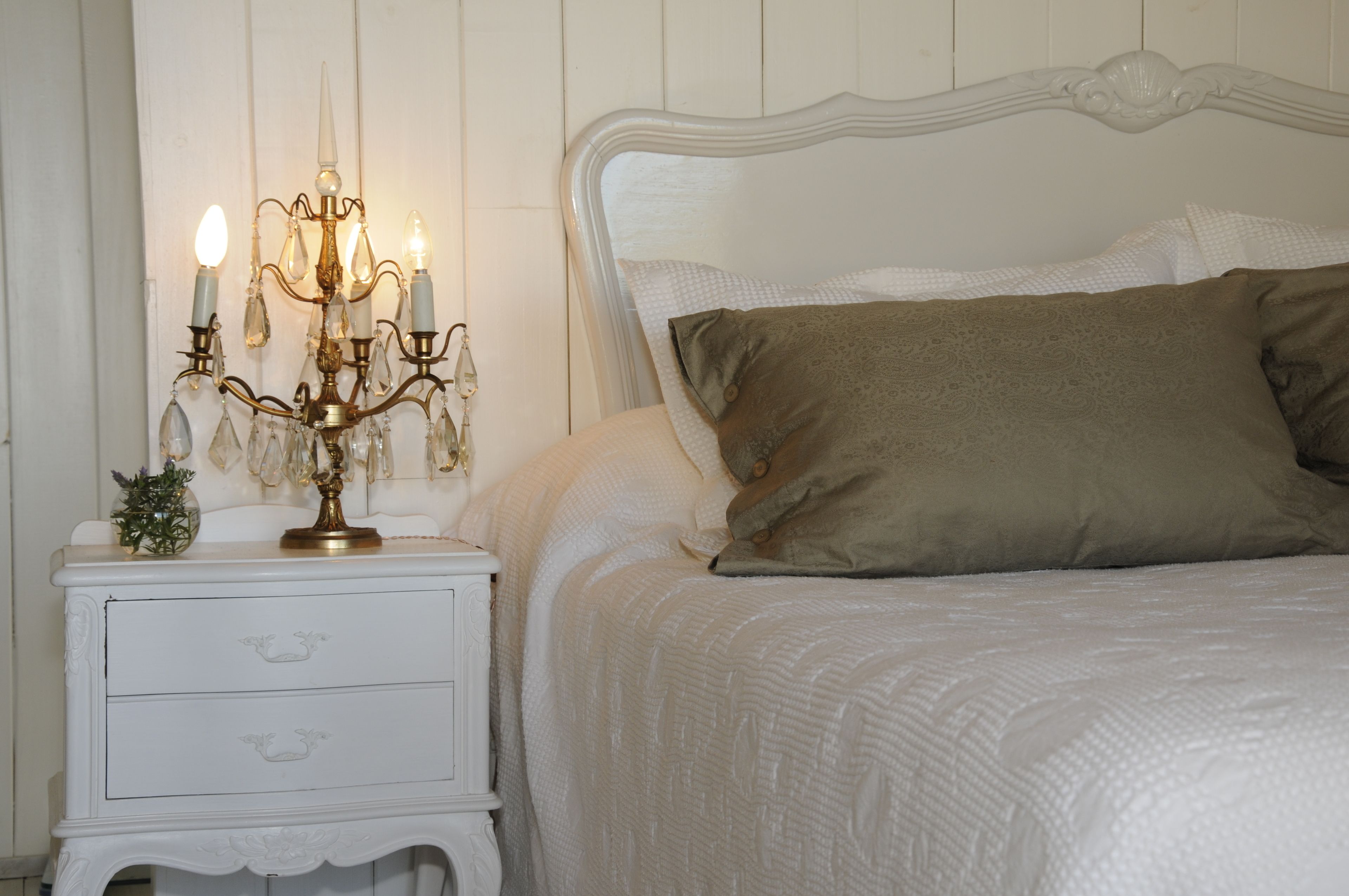 Un lit bien fait, avec une tête de lit blanche, une table de chevet dotée d’un tiroir blanc et un chandelier allumé.