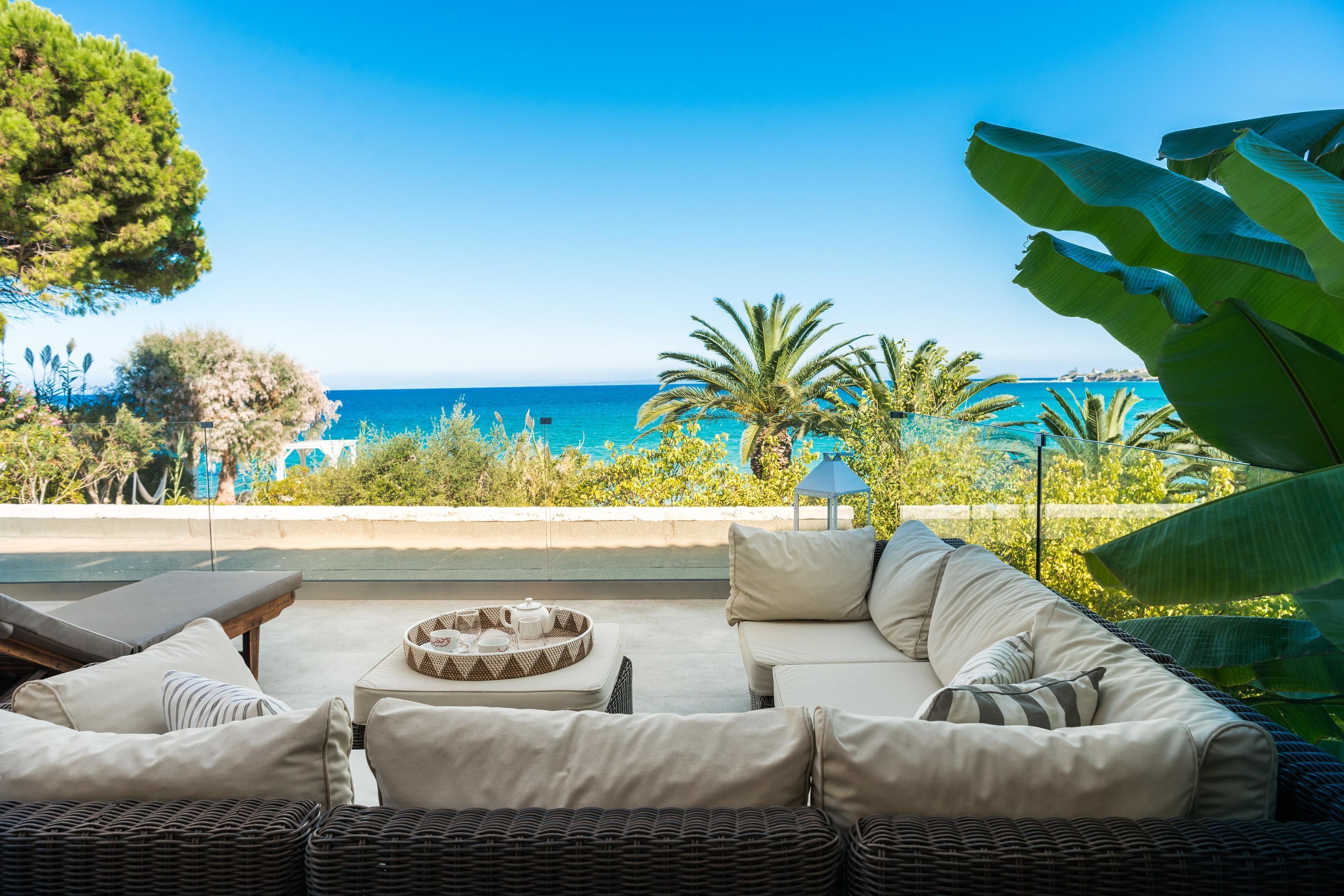A sunlit outdoor seating area with a view of the sea, featuring wicker furniture and a small table with a tray.