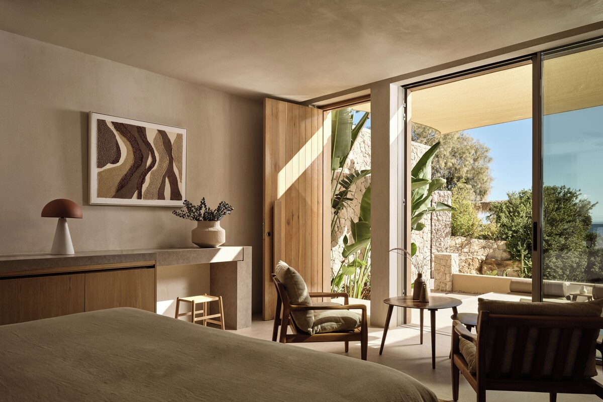 A bedroom with a bed, a chair, a small table, a plant, and a view of the outdoors.