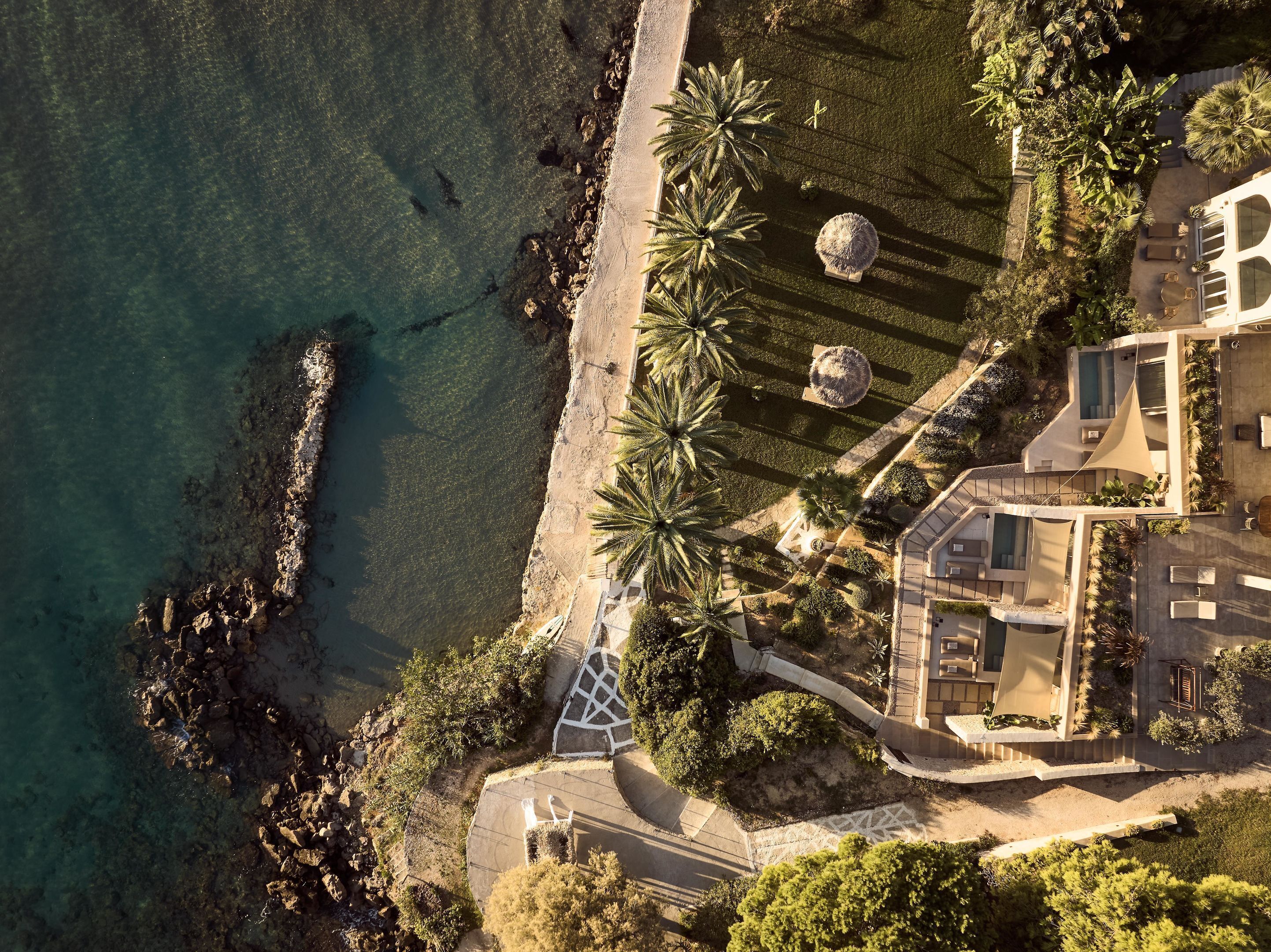 An aerial view of a coastal property with a beach, palm trees, and a modern house with a pool.