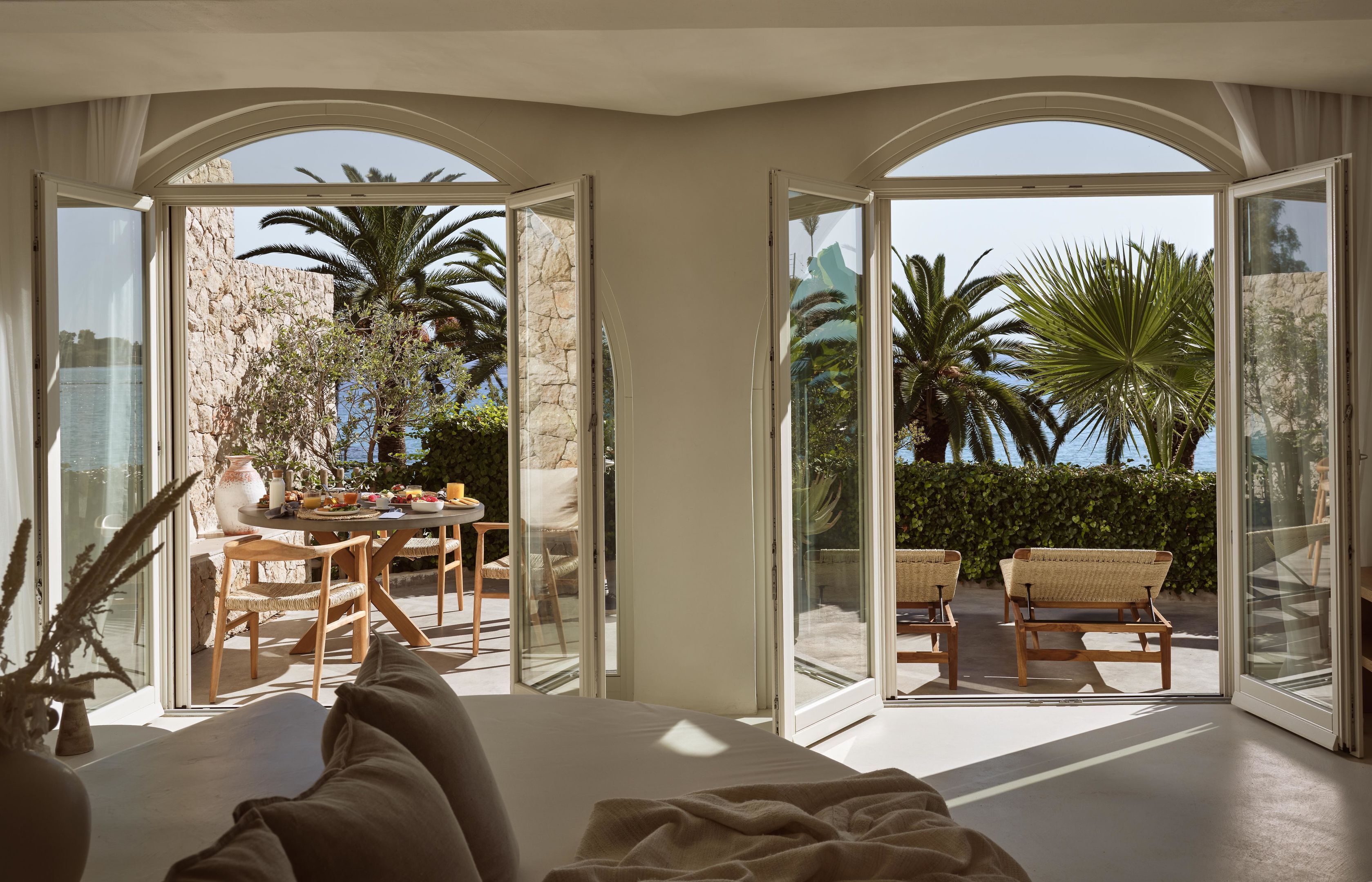 Una habitación con un gran ventanal que da a un espacio exterior con mesa y sillas, rodeado de vegetación y vistas al mar.