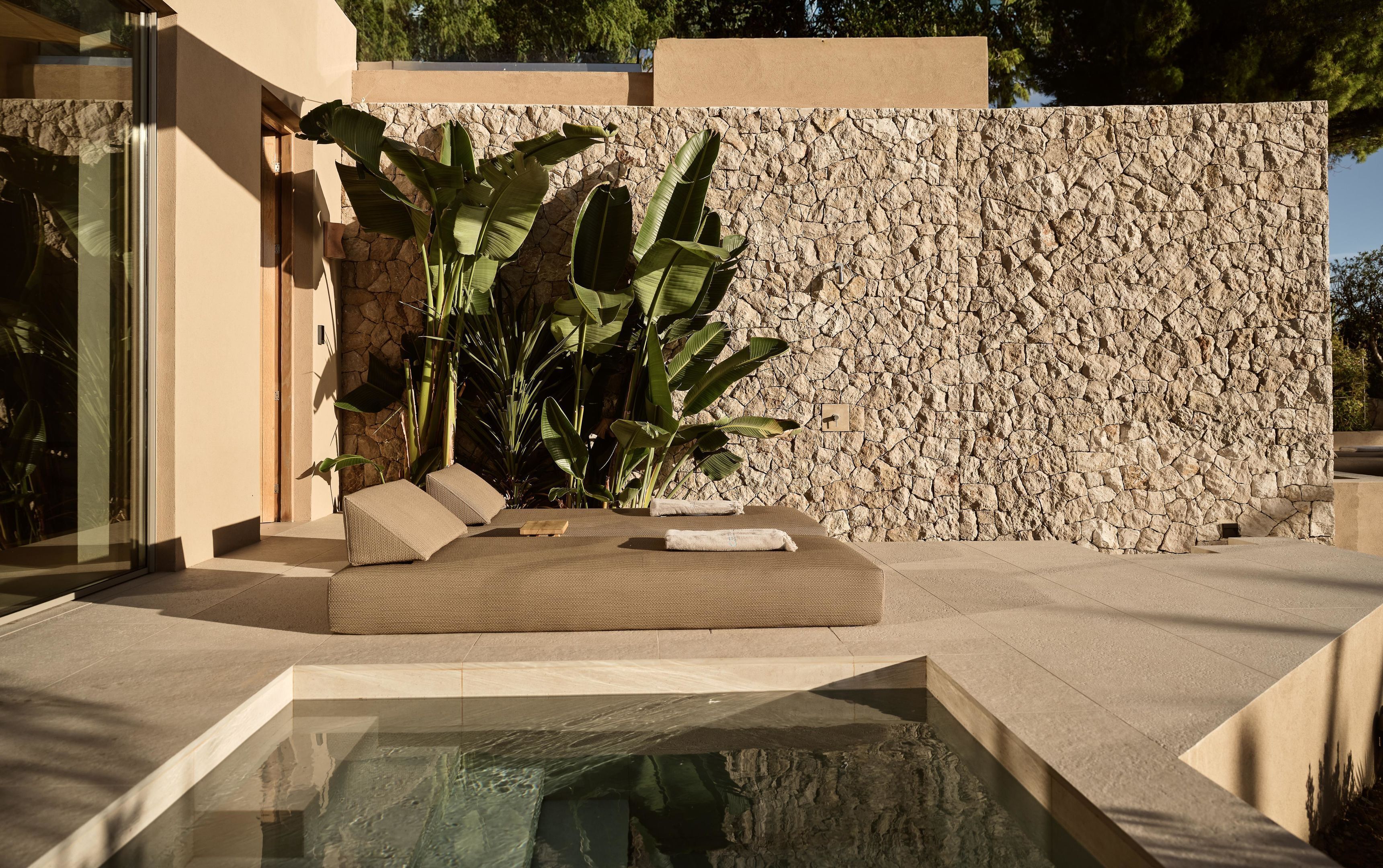 Un espacio moderno al aire libre con un fondo de pared de piedra, una mesa de vidrio y exuberante vegetación tropical.