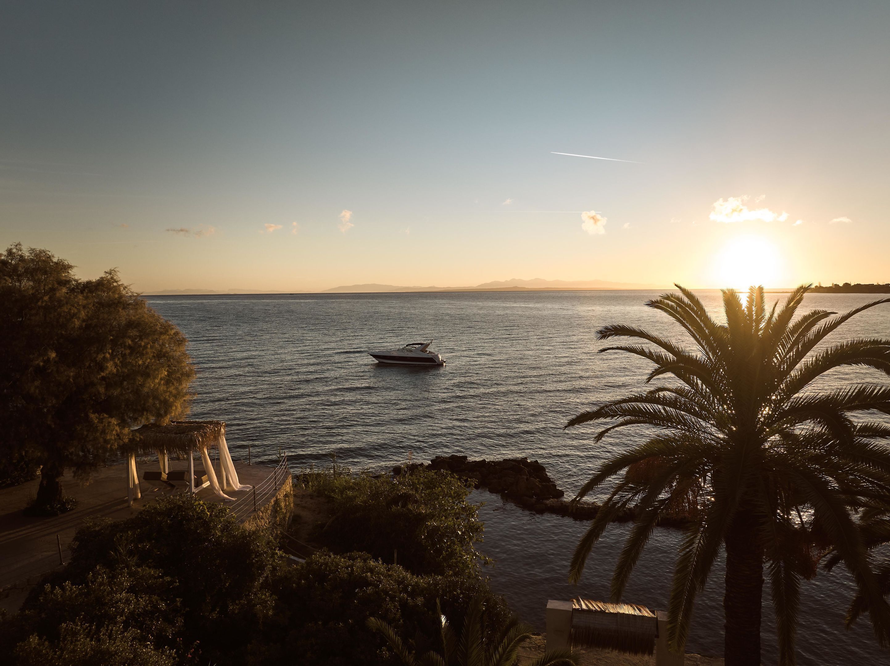 A sunset view over a calm sea with a boat, palm trees, and a rocky shoreline.