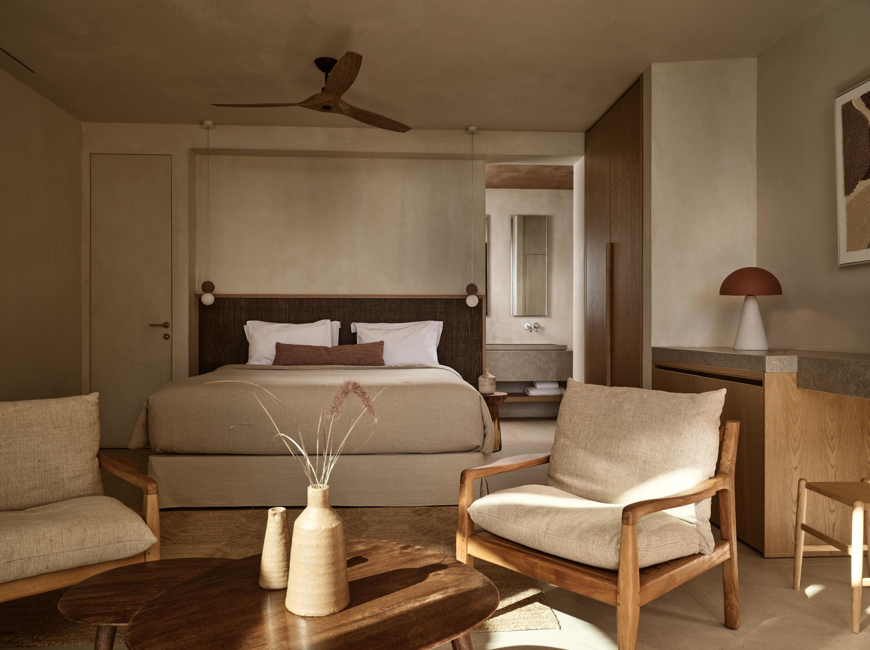 A bedroom with a bed, two chairs, a small table, a nightstand, a ceiling fan, a mirror, and a lamp.