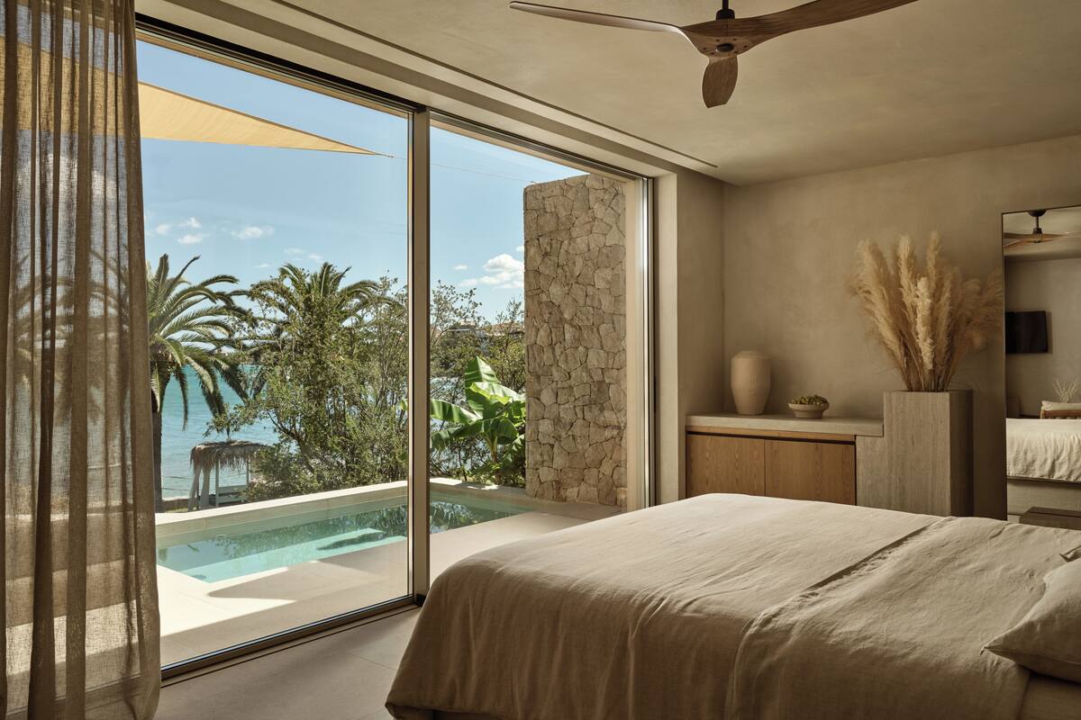 Un dormitorio con un ventanal grande, un ventilador de techo, una cama, una mesita de noche, un florero y vistas a una piscina y zonas verdes.