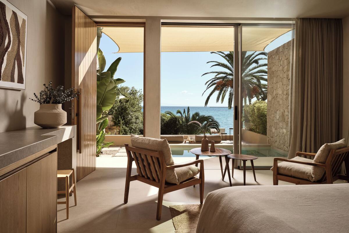 Habitación de hotel con una cama, dos sillas, una mesita, una planta en maceta y vista al mar.