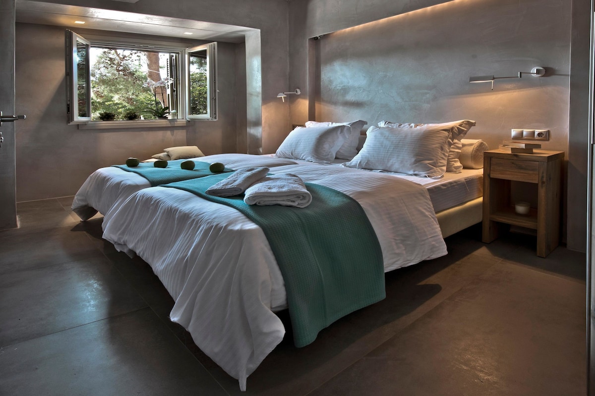 A modern bedroom with a large bed, a bedside table, a window with a view of trees, and a wall-mounted light fixture.