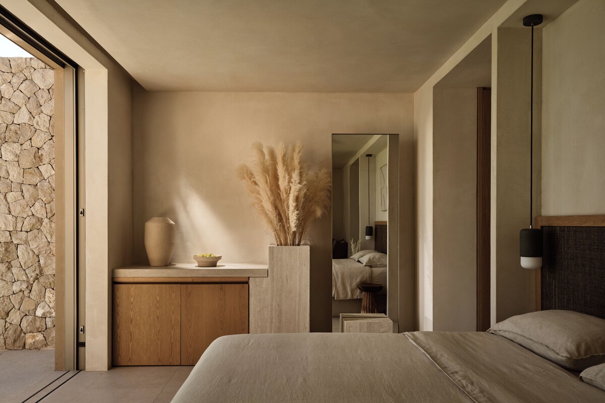 A modern bedroom with a stone wall, a wooden cabinet, a large bed, a mirror, and a vase with pampas grass.