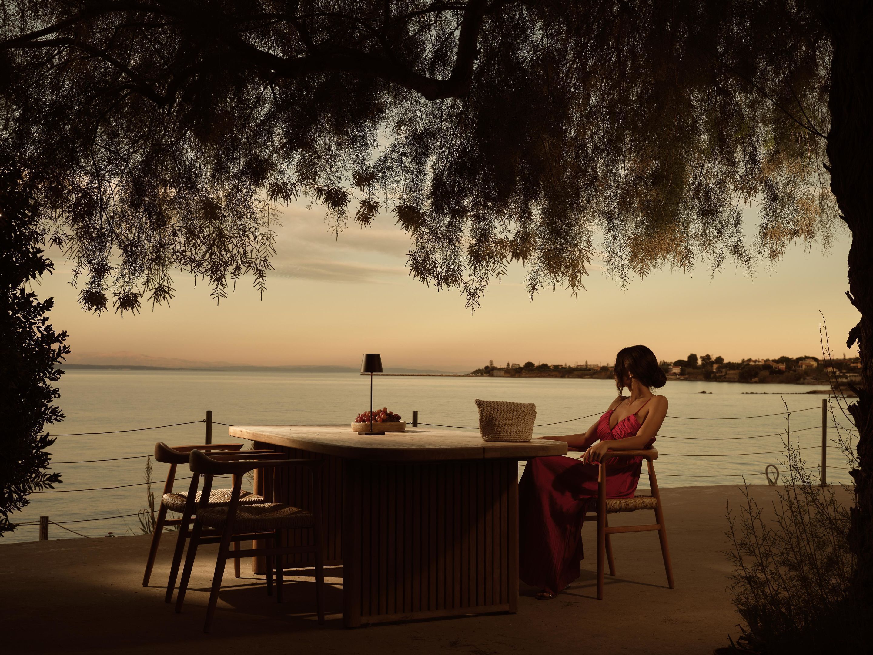 Una mujer sentada en una mesa de madera con una lámpara y una cesta de fruta, contemplando un cuerpo de agua con vistas a una costa lejana.