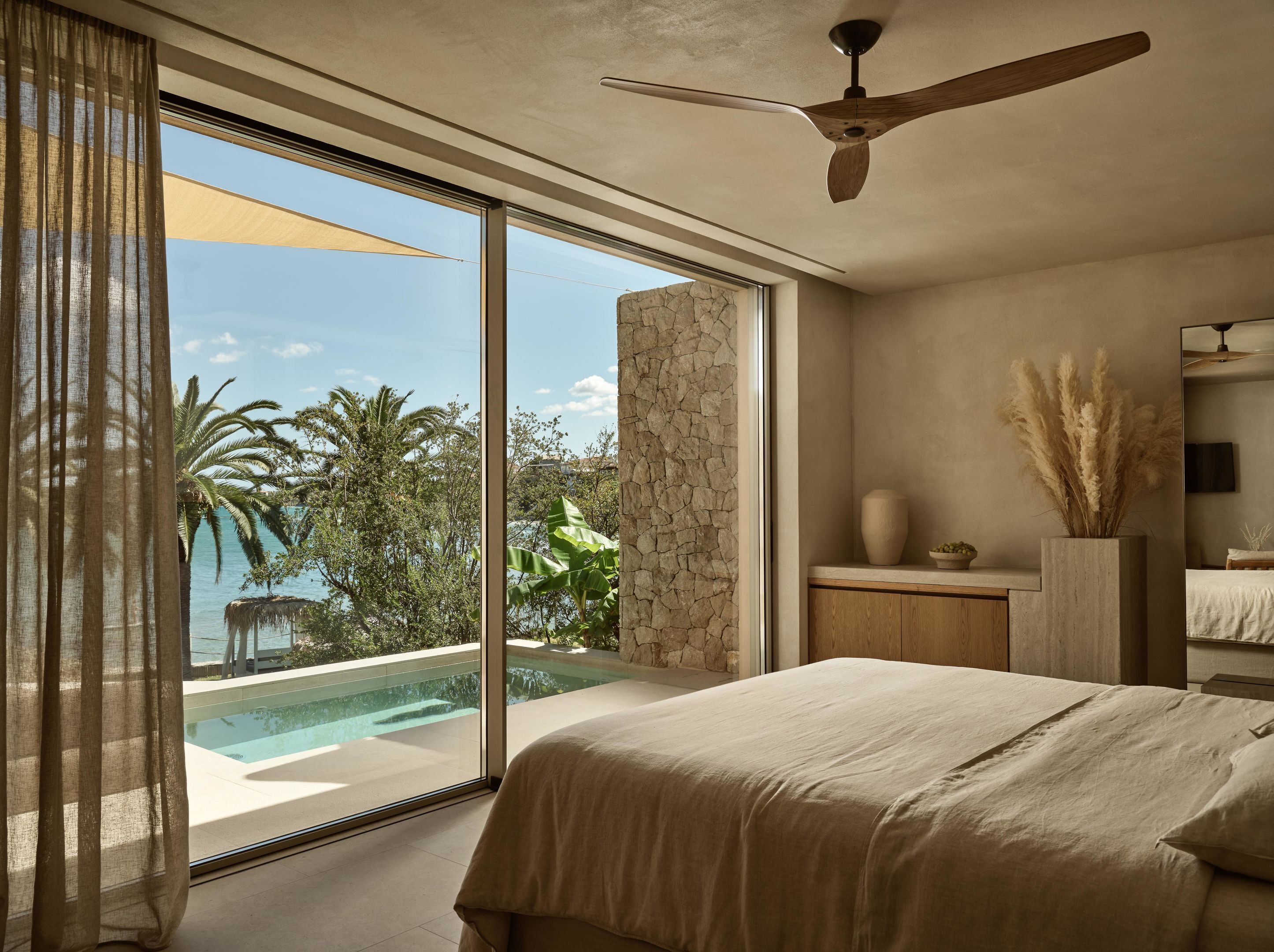 A bedroom with a large bed, a ceiling fan, a stone wall, a small table with a vase, and a view of a swimming pool and palm trees.
