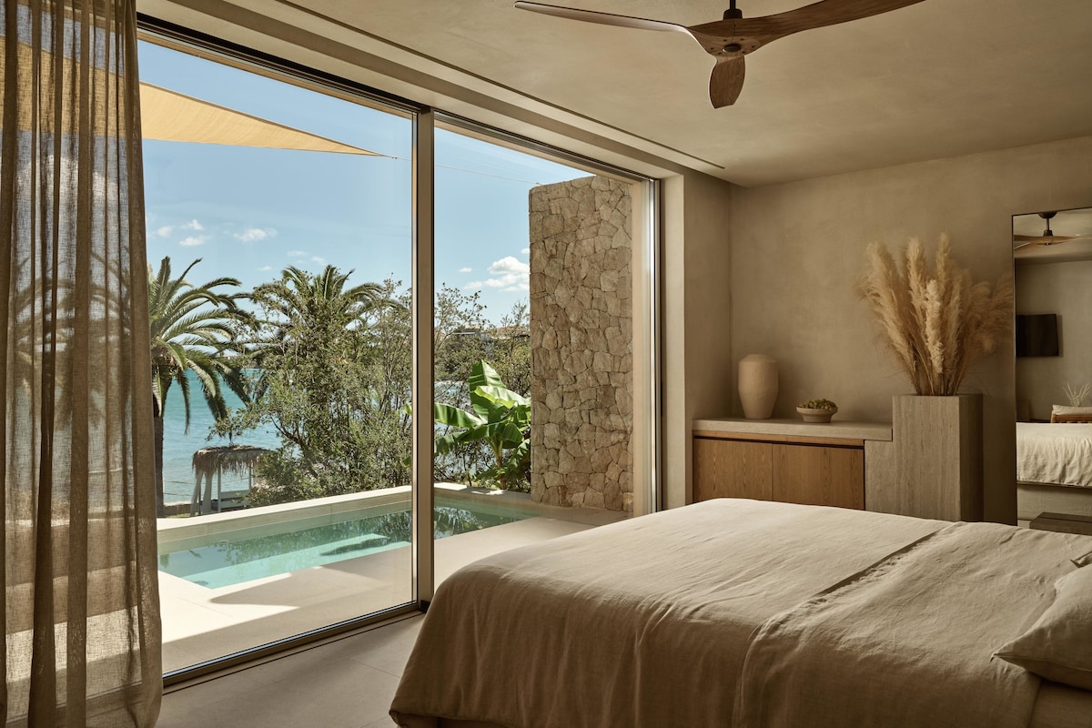 A bedroom with a large bed, a ceiling fan, a stone wall, a small table with a vase, and a view of a swimming pool and palm trees.