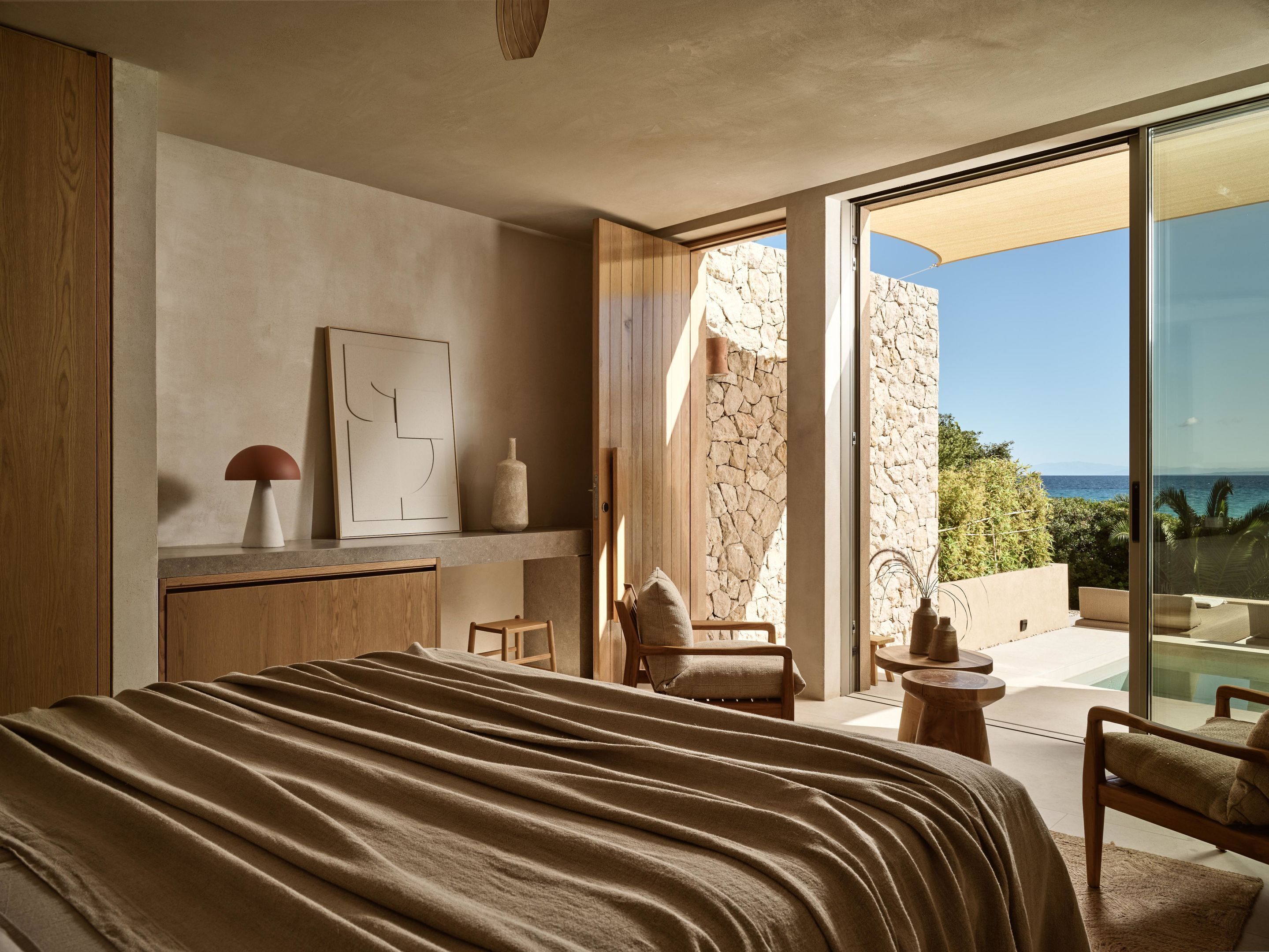 Quarto moderno com uma cama grande, cômoda de madeira, parede de pedra e varanda com vista para o oceano.
