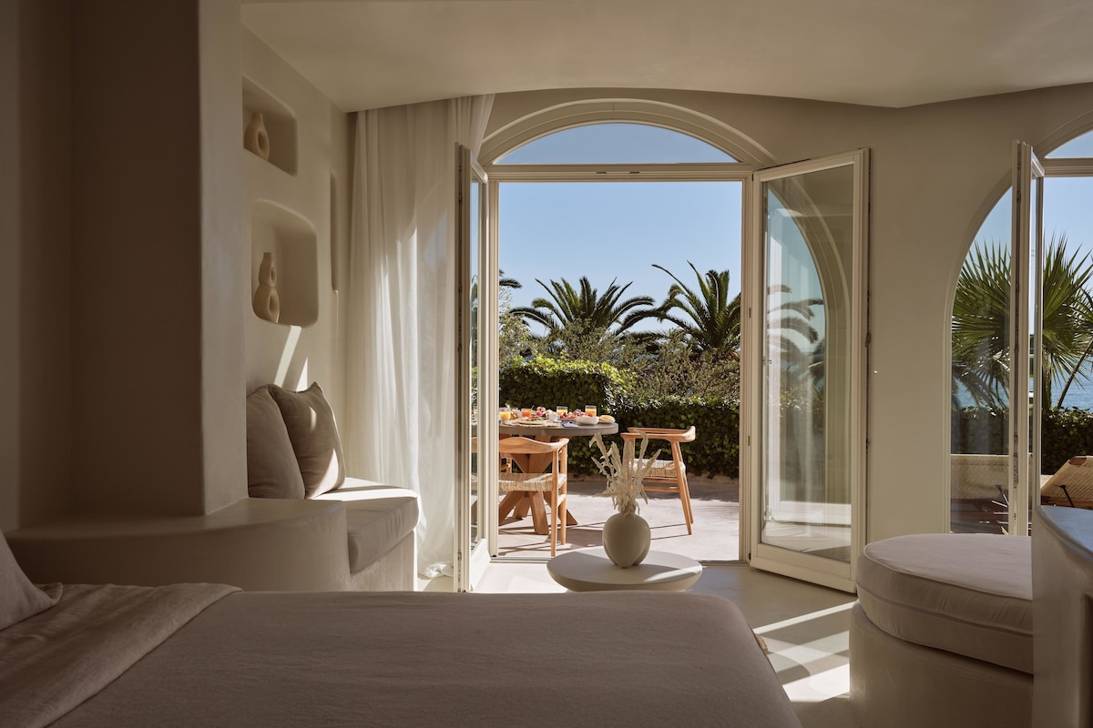 A modern bedroom with a large bed, a sitting area with a table and chairs, and a view of the outdoors through a glass door.