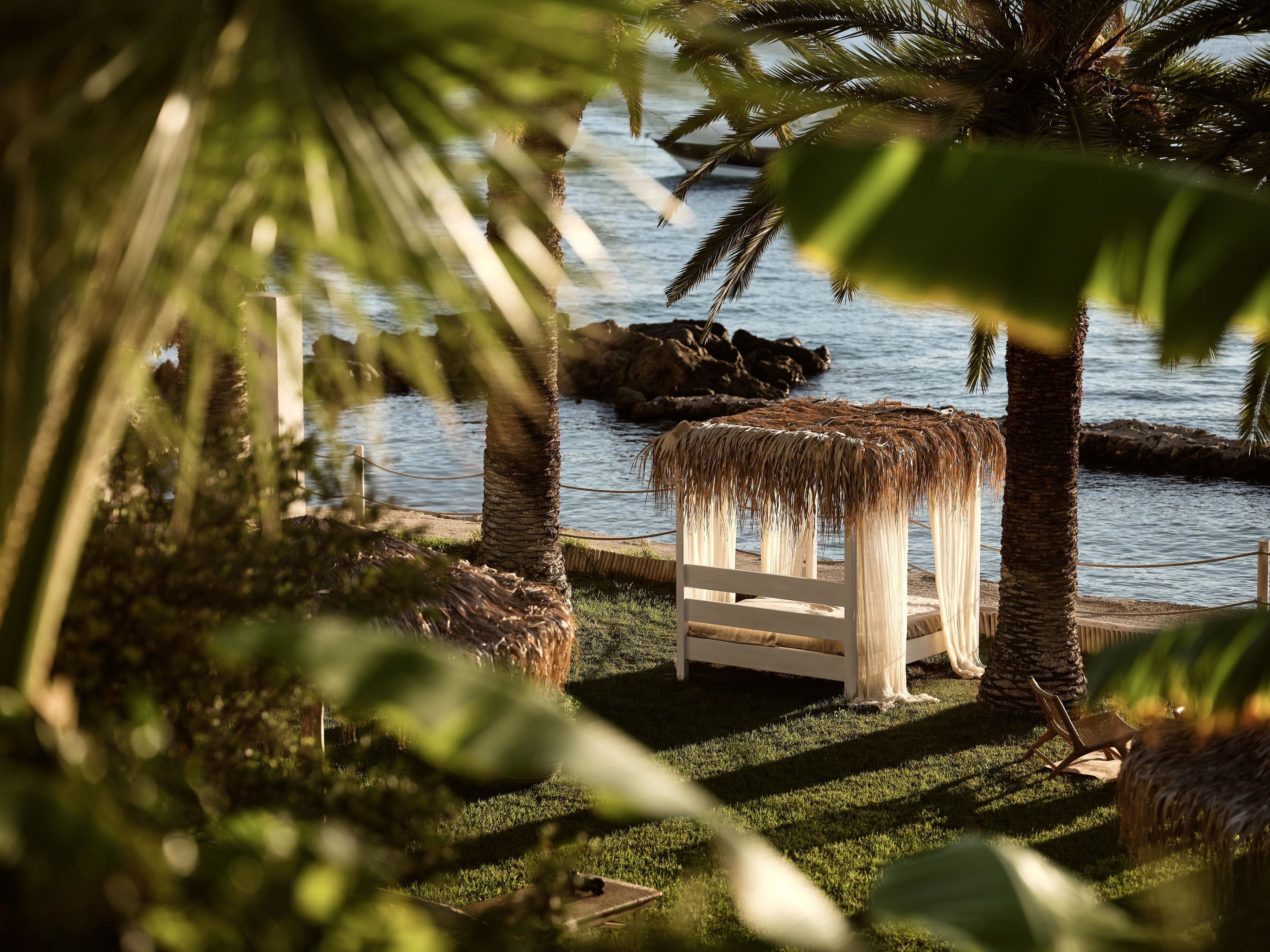 A beachside seating area with a thatched roof, surrounded by palm trees and a view of the sea.