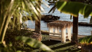 A beachside seating area with a thatched roof, surrounded by palm trees and a view of the sea.