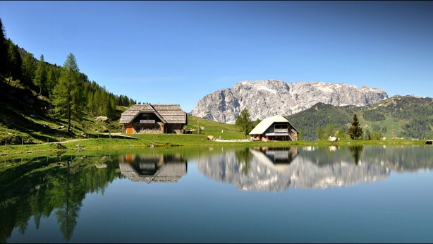 A serene lakeside scene with two rustic cabins, one with a sloped roof and the other with a flat roof, surrounded by lush greenery and a mountain backdrop. - Enzianbrenner Bed & Breakfast (Hermagor-Pressegger See)