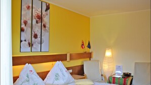 A hotel room with two beds, floral bedding, a wooden headboard, a bedside table with a lamp, and a wall with two framed pictures and two flags. - Enzianbrenner Bed & Breakfast (Hermagor-Pressegger See)